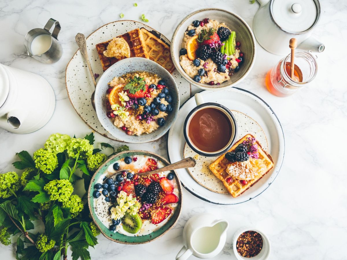 The image shows a breakfast setting with bowls of oatmeal, waffles with fruit, syrup, coffee, milk, and fresh greenery on a white marble surface.