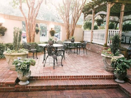 Outdoor patio with wrought-iron furniture, brick flooring, large potted plants, shaded by trees and a wooden pergola, inviting and serene atmosphere.