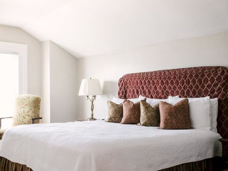 A bedroom with a large bed featuring a red patterned headboard, multiple brown and white pillows, a bedside lamp, and a chair by the window.