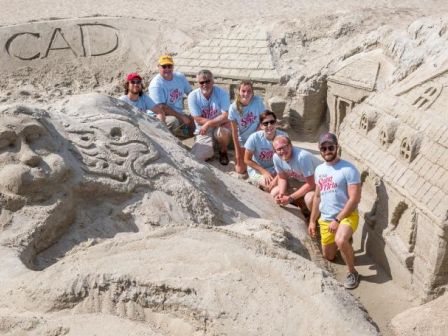 A group of people in matching shirts pose in front of intricate sand sculptures on a beach, with 