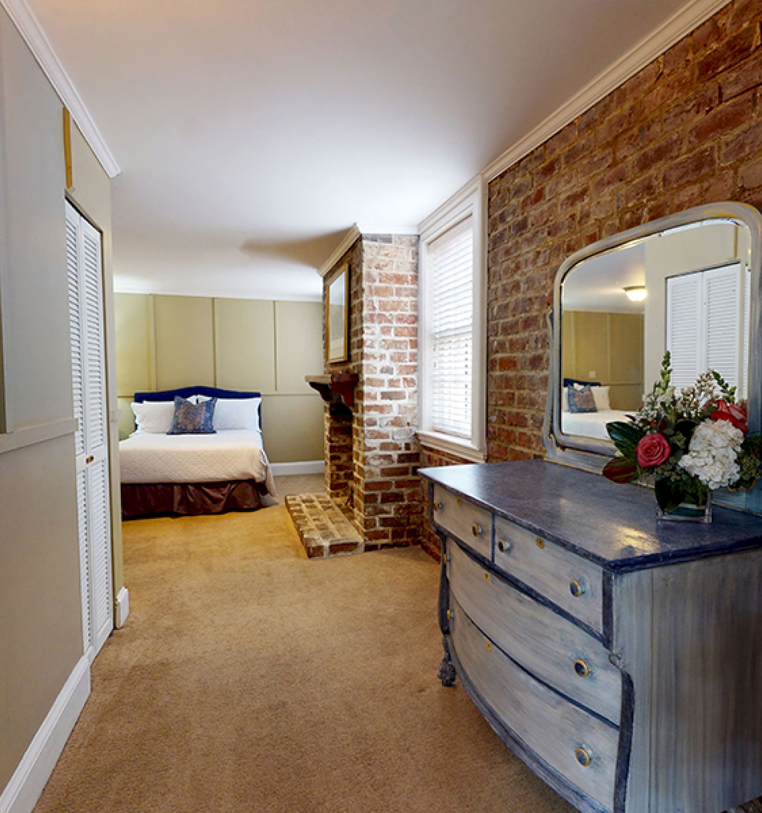 A cozy bedroom with a brick accent wall, a bed with blue and white bedding, a vintage dresser with a mirror and flowers, and carpeted floor ends the sentence.