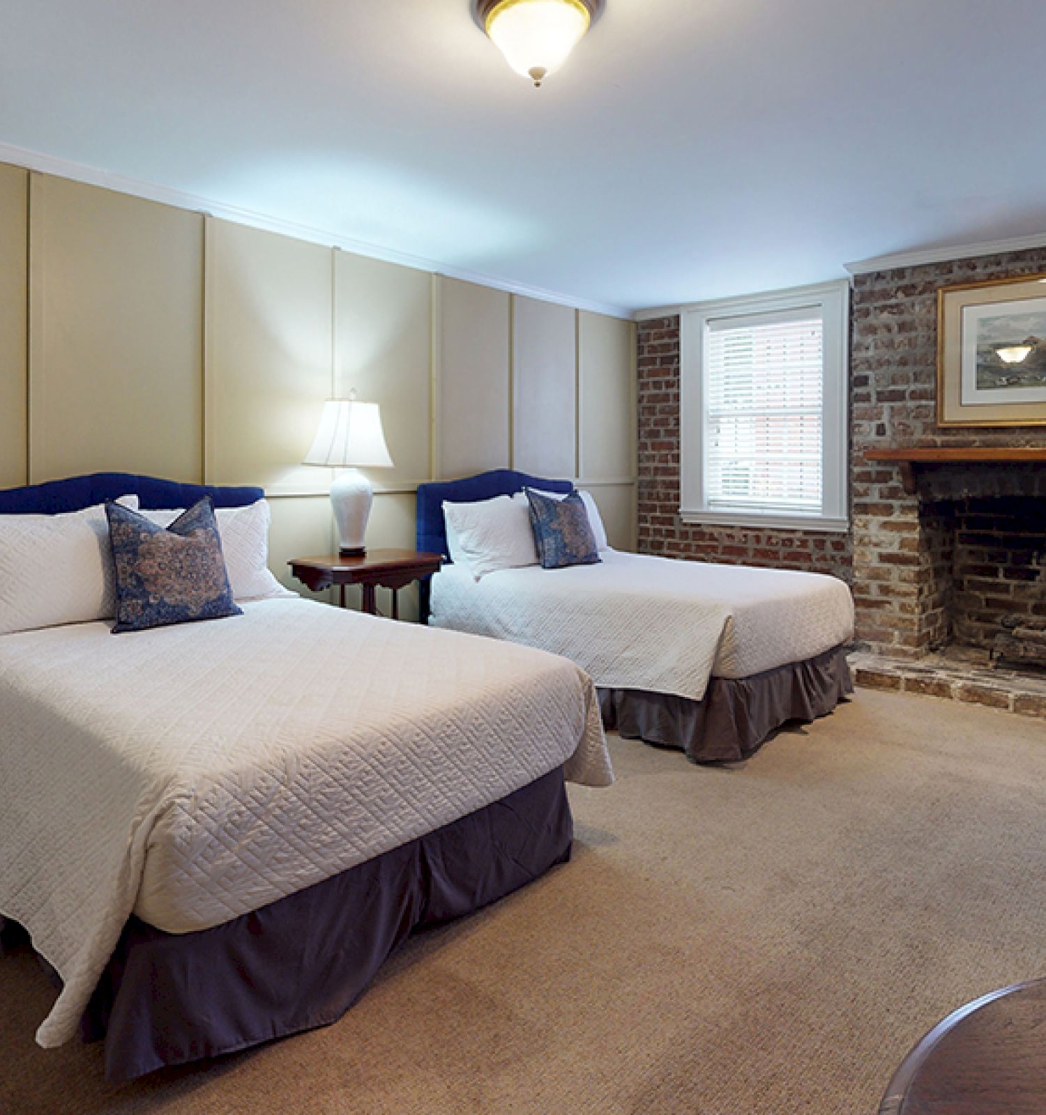 The image shows a cozy bedroom with two double beds, a nightstand with a lamp, and a brick fireplace, creating a warm, inviting atmosphere.