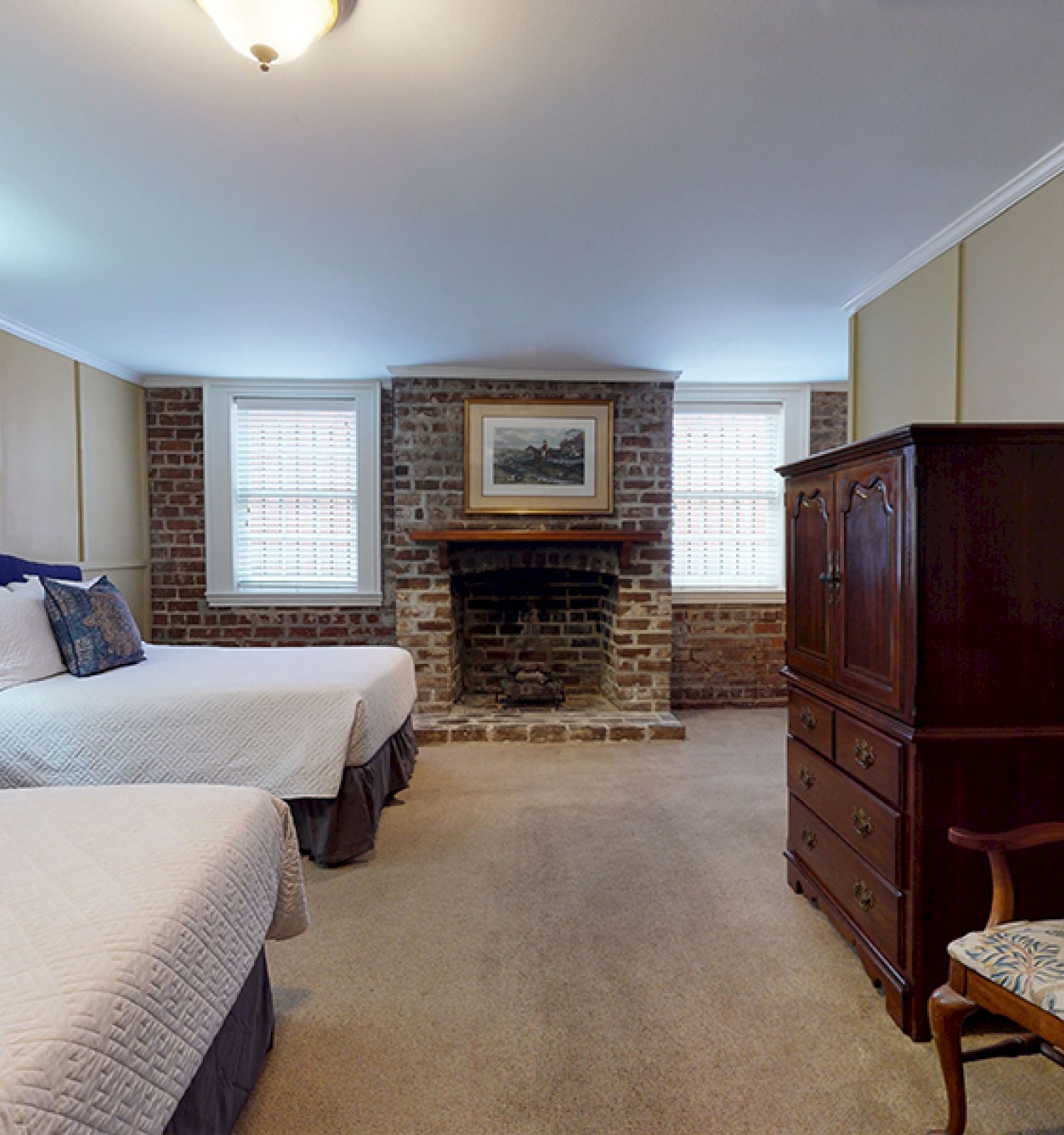 A cozy bedroom with two double beds, a wood dresser, a chair, a small table, and a brick fireplace between two windows.