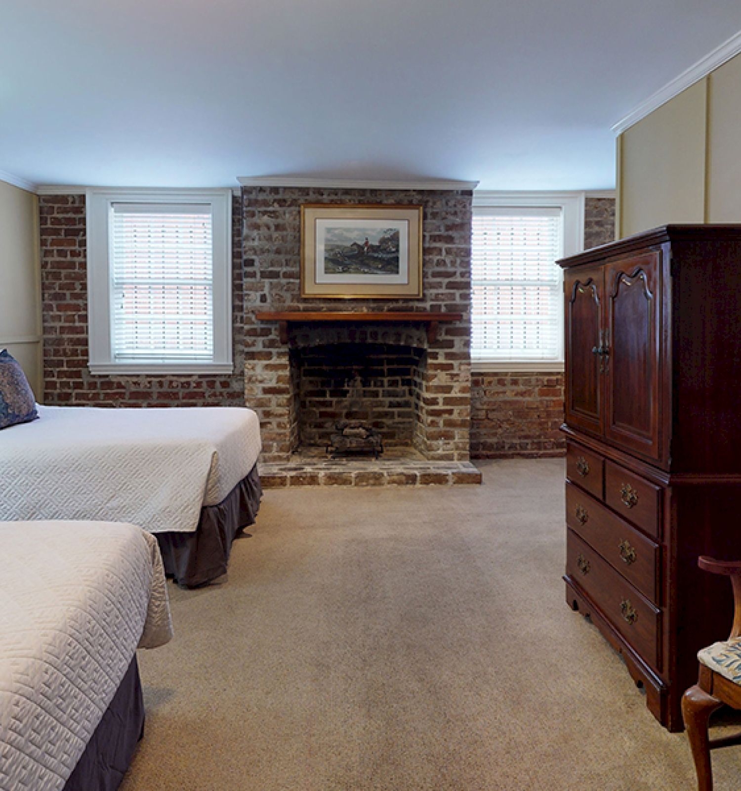 The image shows a cozy bedroom with two beds, a fireplace between two windows, an antique wardrobe, a wooden chair, and a painting above the fireplace.