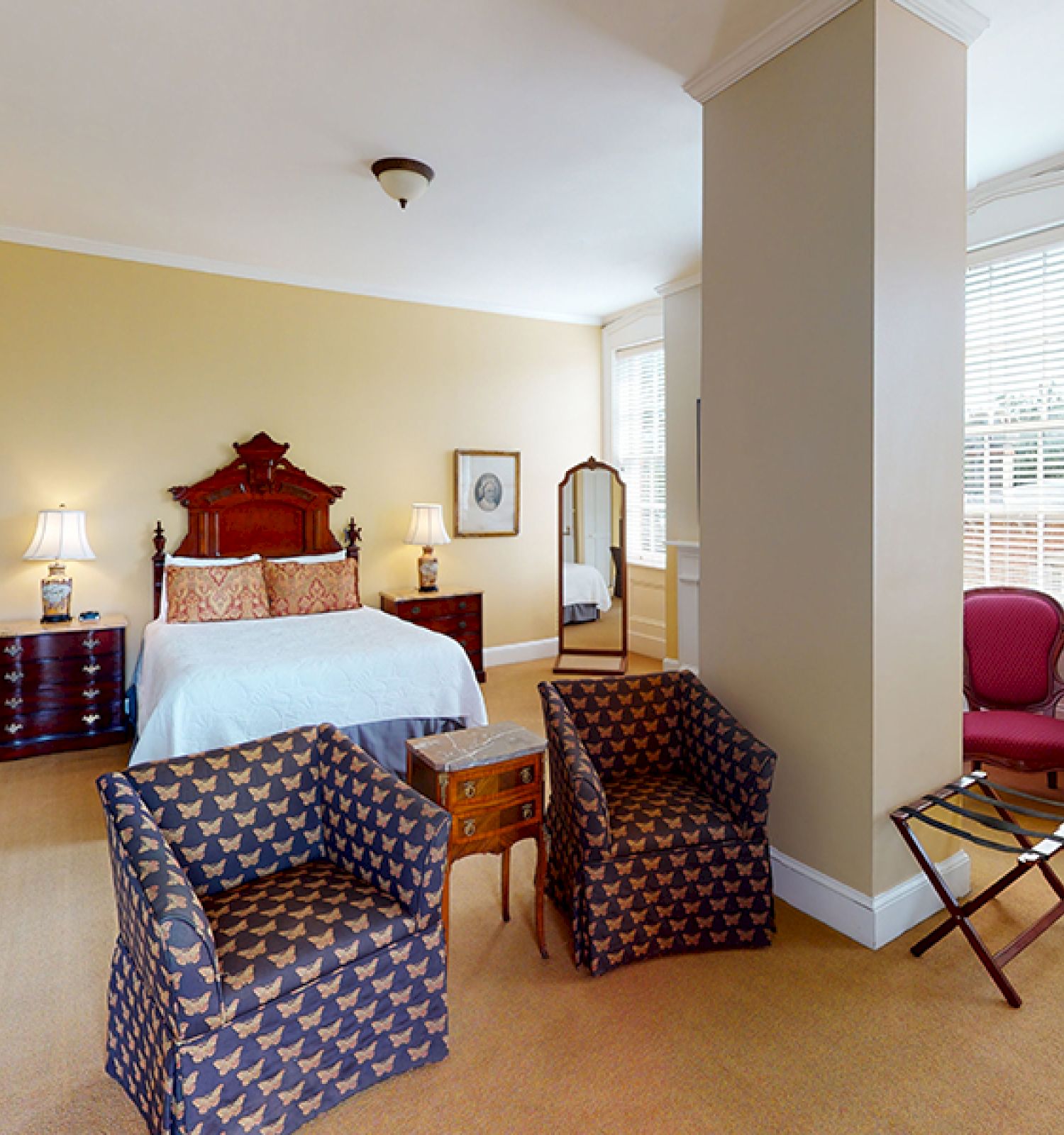 A spacious bedroom features a large bed, two patterned chairs, a dresser, a mirror, and a small table with a chair near a window.