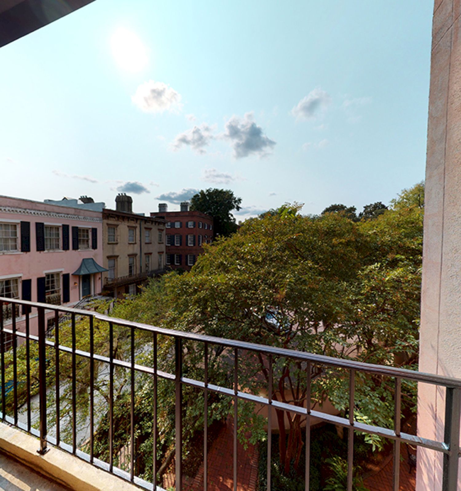 A small balcony overlooks a lush green courtyard and nearby buildings under a sunny, partly cloudy sky.