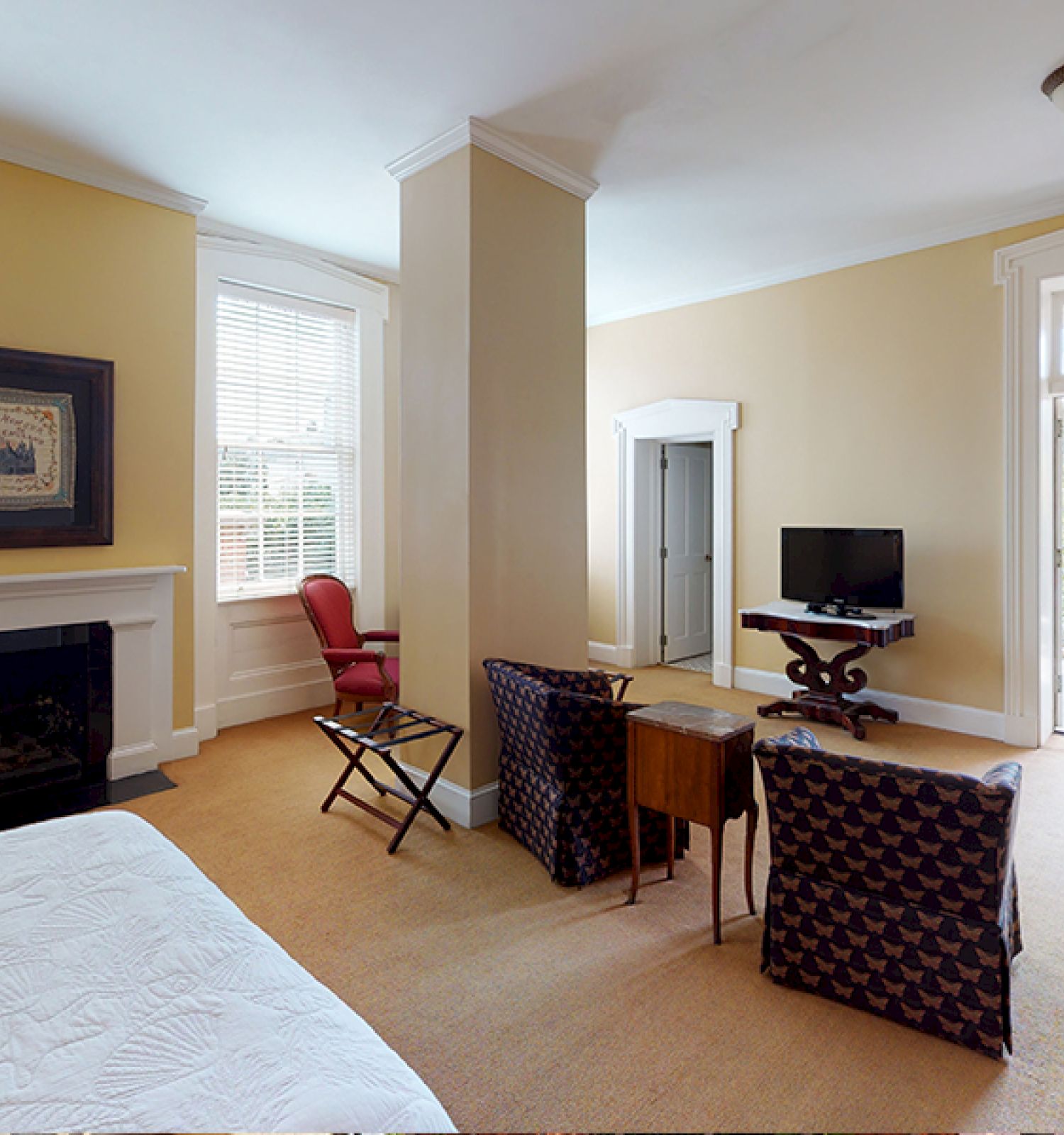 A spacious room with a bed, chairs, a TV, a fireplace, and a balcony door, featuring yellow walls and large windows, and a red chair.