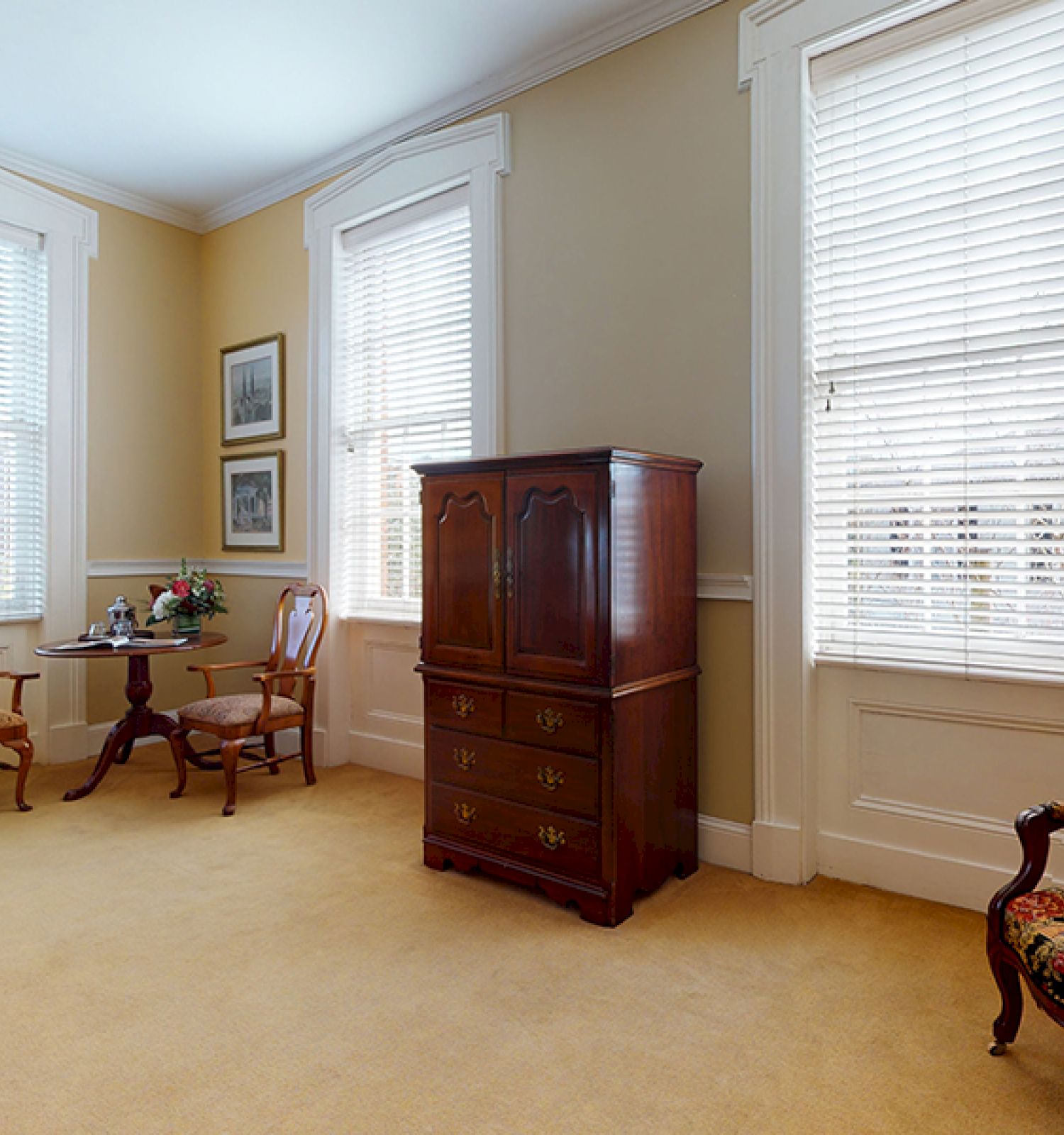 The image shows a cozy room with antique furniture, including a wooden cabinet, a small table with two chairs, and a patterned armchair.