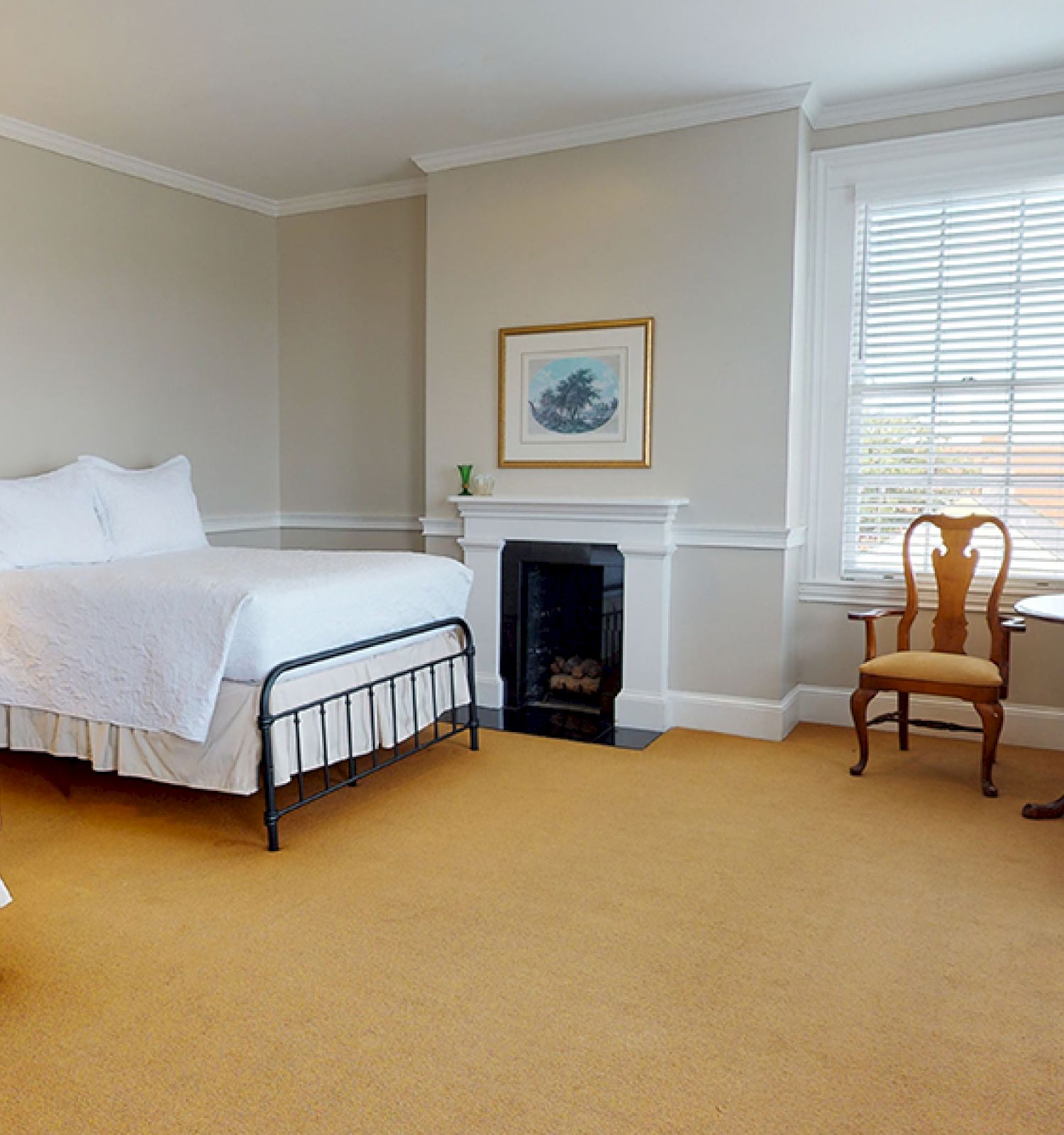 A tidy bedroom features two beds, a fireplace, a window, a table with chairs, and a wooden dresser, all in a neutral color scheme.