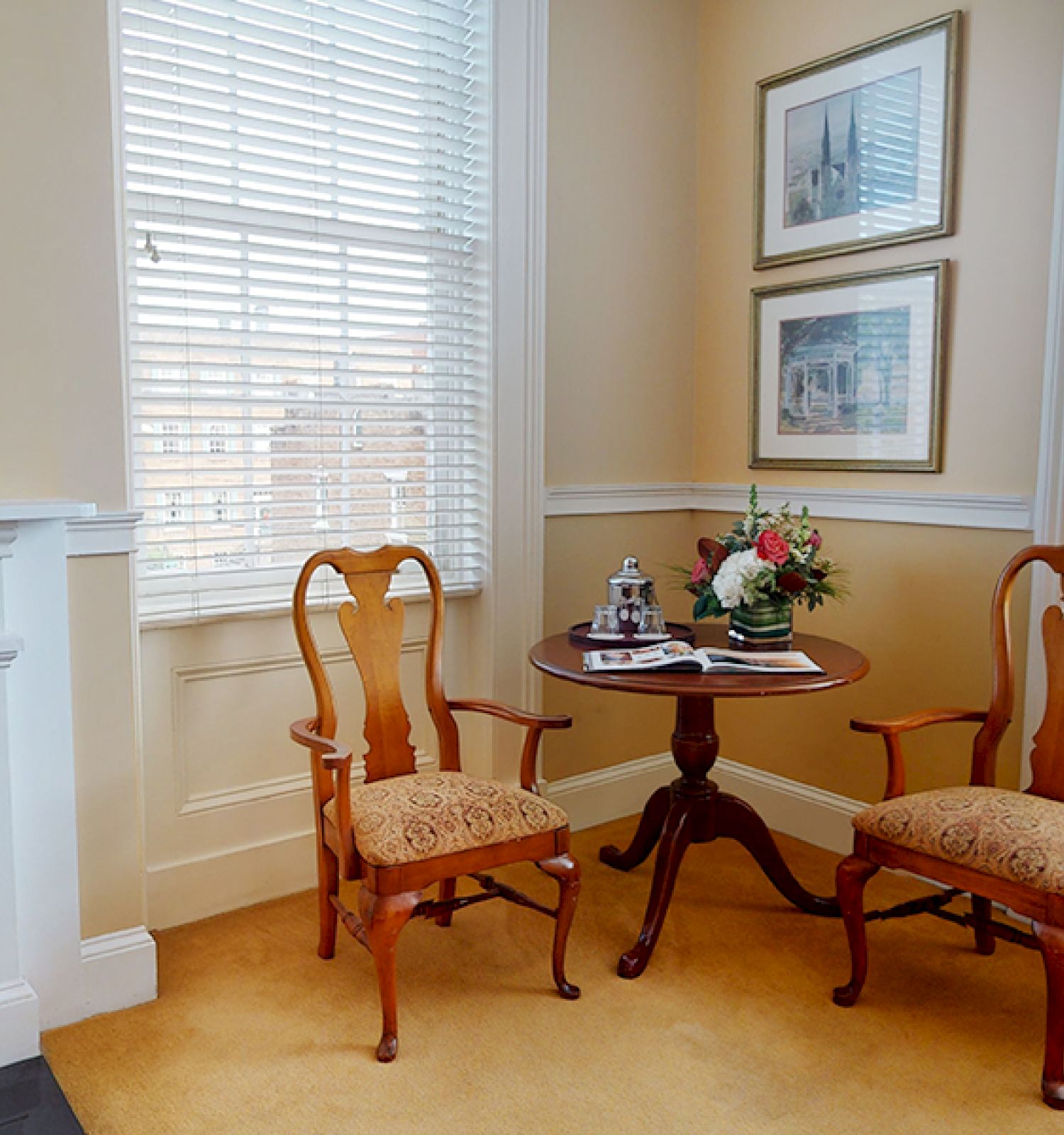 A cozy room with two wooden chairs around a small round table, flowers, books, and framed pictures on the wall, white blinds on the windows.