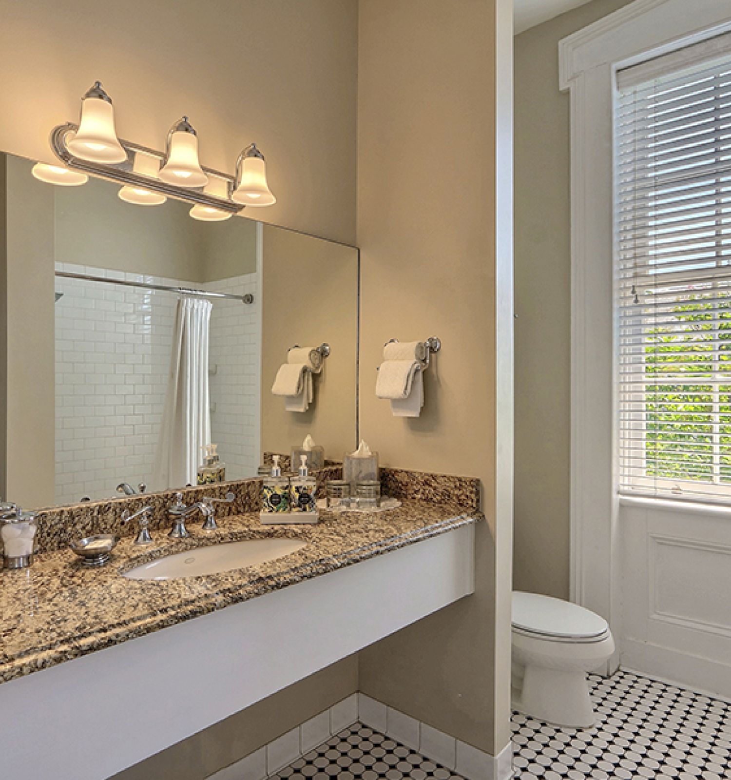 The image shows a bathroom with a granite countertop sink, a toilet, a window with blinds, and a tiled floor. A shower is visible through an open door.