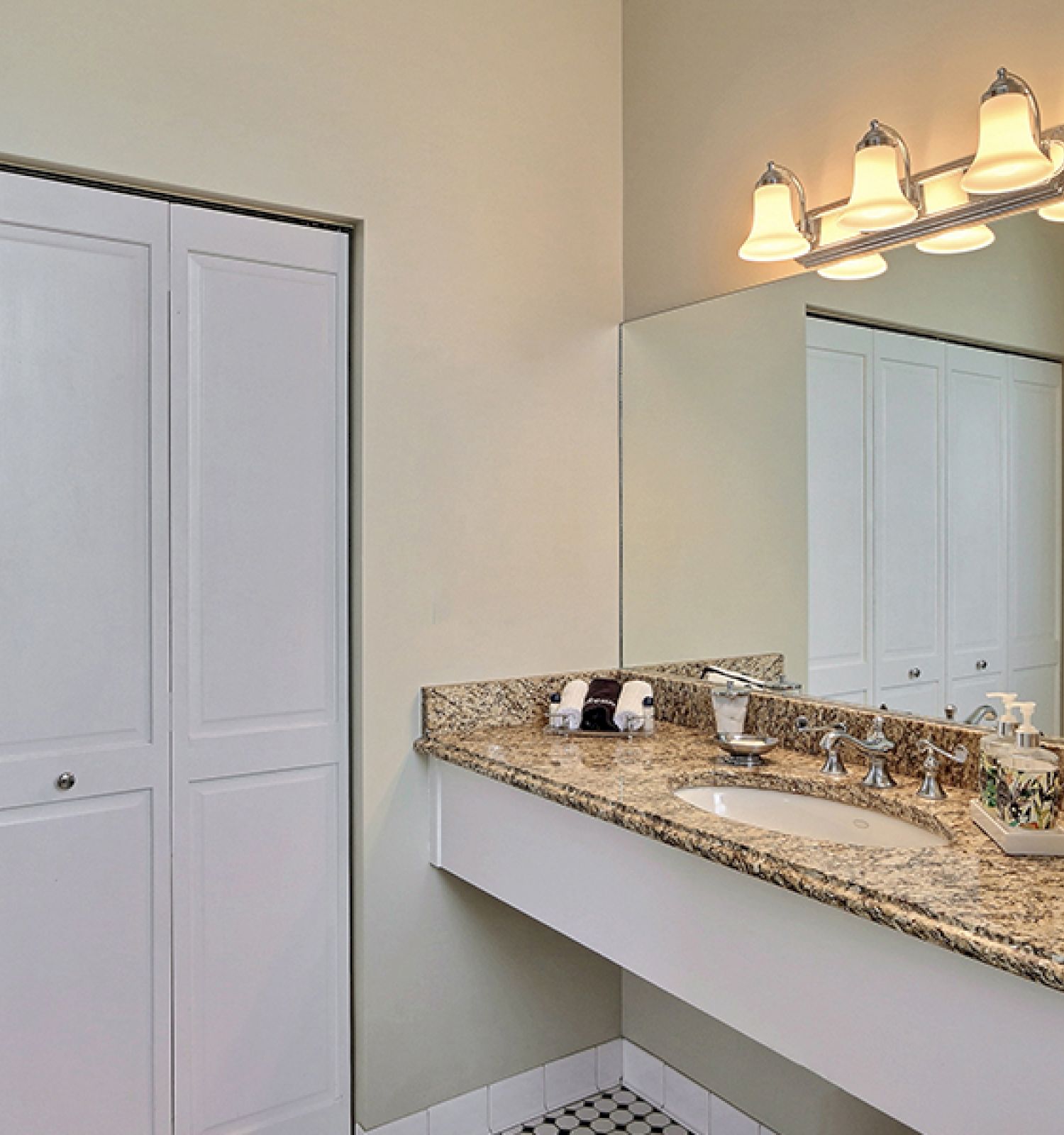 A bathroom with granite countertops, a large mirror, under-sink storage, a closet with white doors, and a light fixture with four lights.