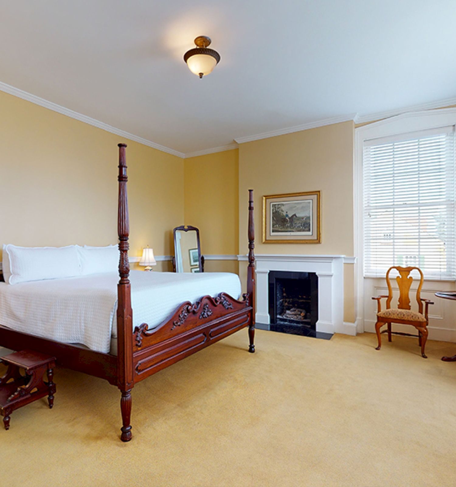 A bedroom with a four-poster bed, nightstands, a chair, a desk with flowers, window shades, a fireplace, a mirror, and a lamp on the ceiling.