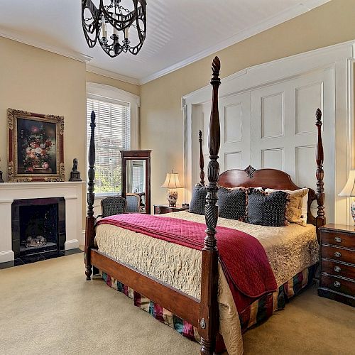 A cozy bedroom with a four-poster bed, decorative pillows, a fireplace, wall art, bedside tables with lamps, and a chandelier overhead.