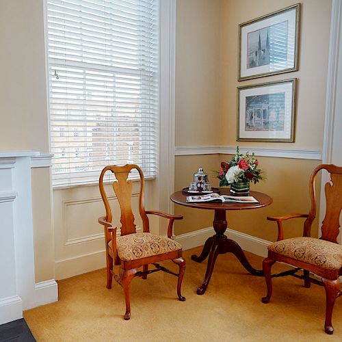 The image shows a small seating area with two wooden chairs and a round table, decorated with flowers and magazines, near two windows.