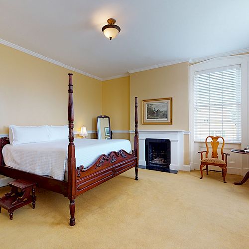 A bedroom with a wooden four-poster bed, nightstands, chairs, a fireplace, a mirror, a window with blinds, and a small desk with a flower vase.