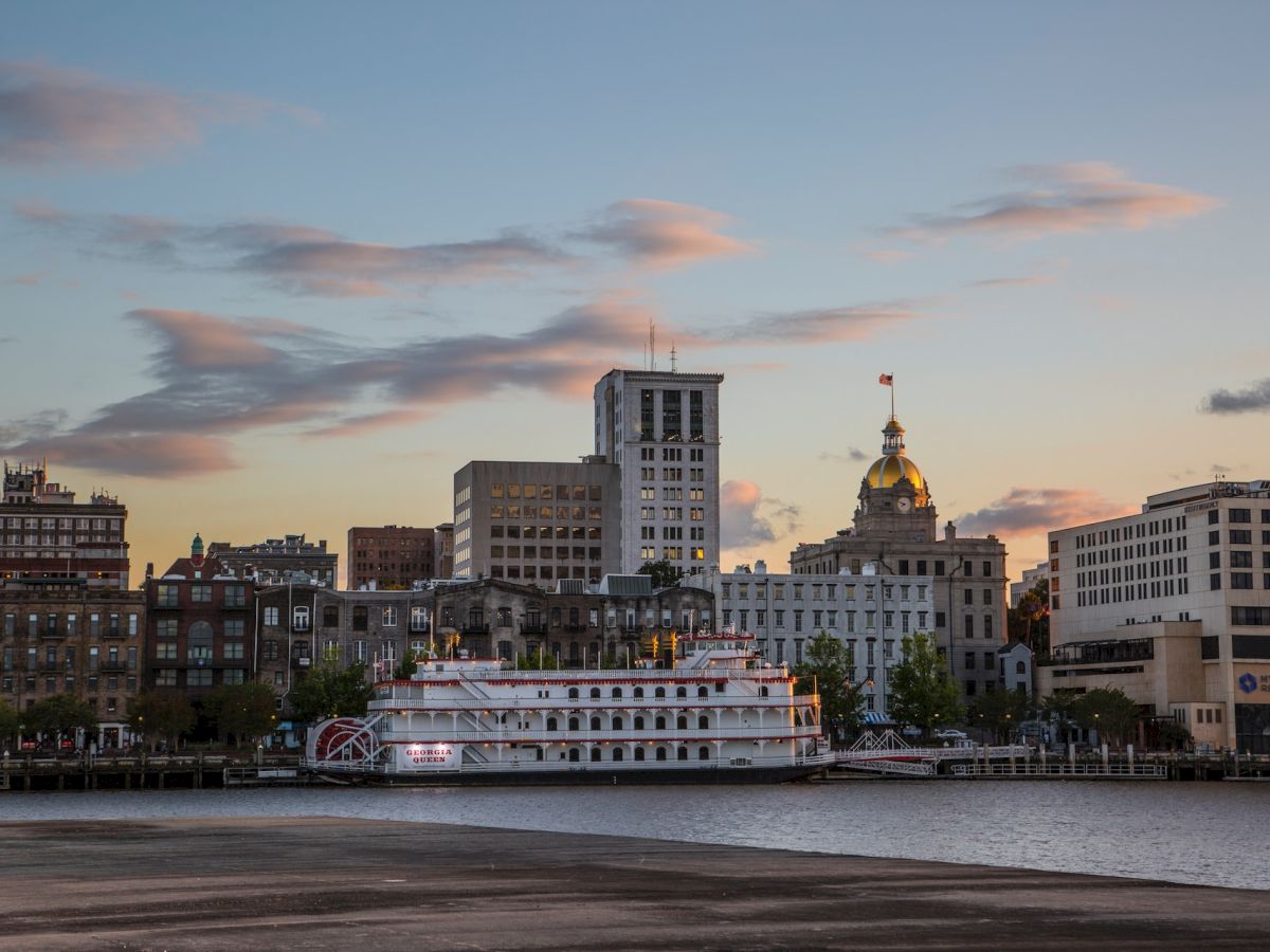 A scenic cityscape with a river, a docked riverboat, and various buildings, including a domed structure, against a sunset sky.