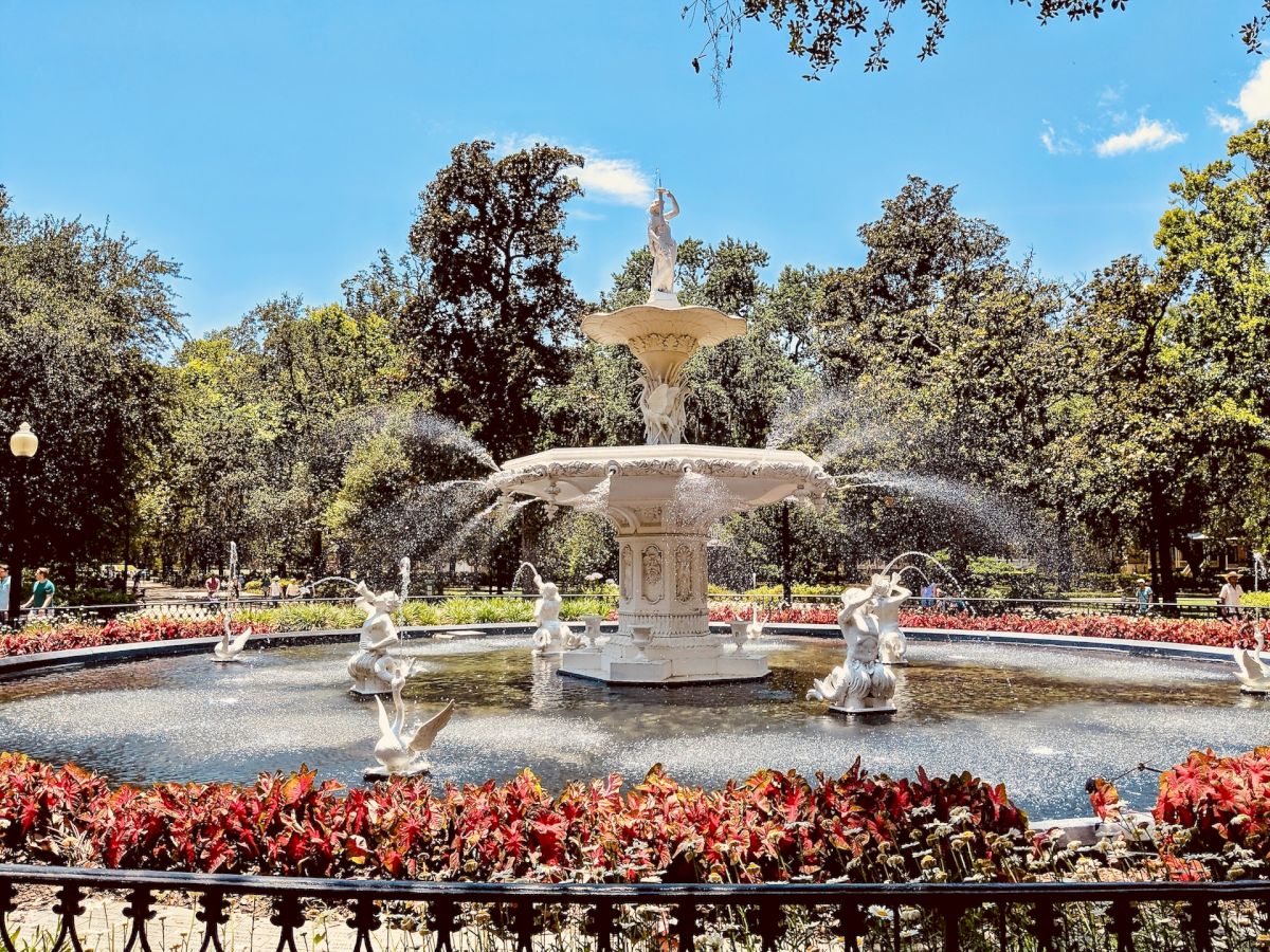 A white, multi-tiered fountain surrounded by water, statues, and red flowers is in a park with green trees and a bright blue sky.