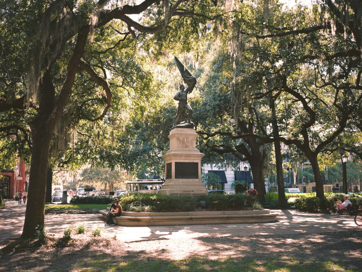 A serene park scene with a statue at the center, surrounded by large trees and scattered visitors relaxing on benches or walking.