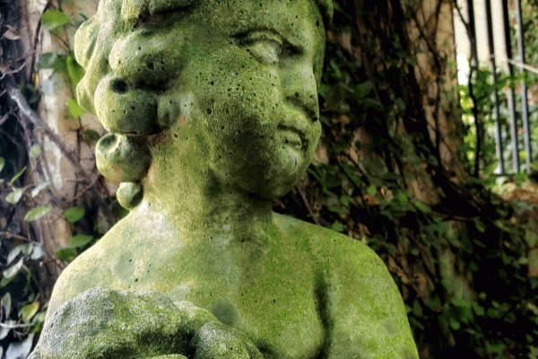 The image shows a moss-covered stone statue of a child holding a cluster of fruit, surrounded by ivy and greenery.