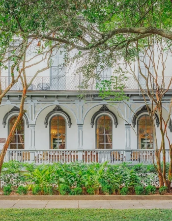 A charming white building with arched windows and intricate ironwork, framed by trees and lush greenery at the front.