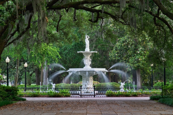 A fountain in a lush park setting, surrounded by trees and greenery, with water jets and decorative statues, under a canopy of branches.