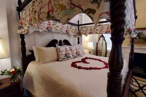 A cozy bedroom with a canopy bed, rose petals arranged in a heart shape on the quilt, and a bouquet on the side table.